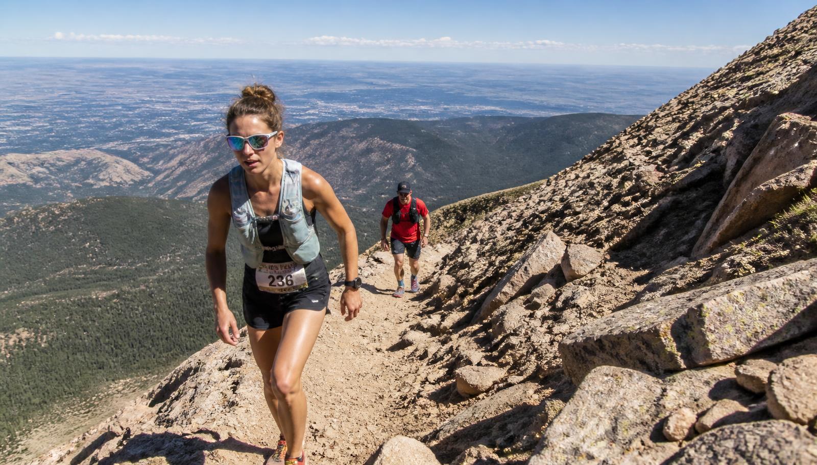Runners climbing a rocky switchback above treeline with the plains far below